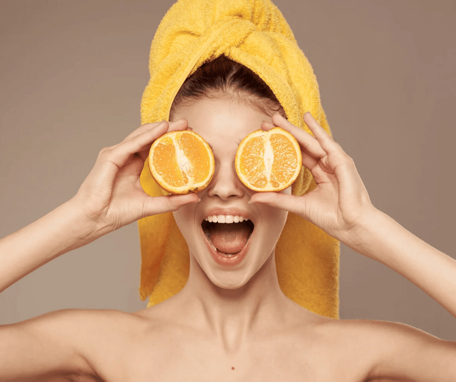 Eine fröhliche Frau hält zwei Orangenhälften vor ihren Augen und hat ein orangefarbenes Handtuch auf dem Kopf eingewickelt.