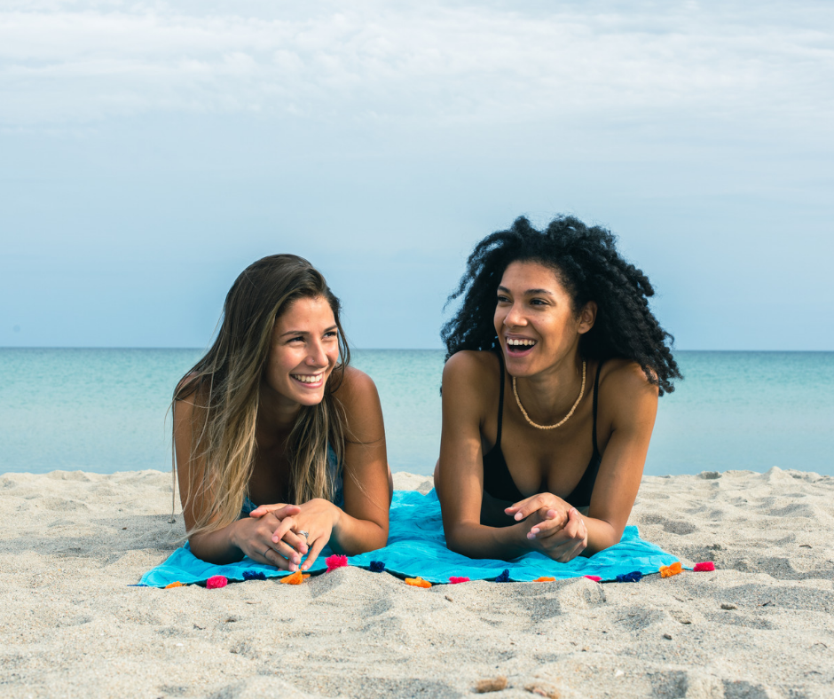 Zwei glückliche Freundinnen, eine weiße und eine farbige, liegen lachend und entspannt auf einem Strandtuch am Sandstrand. Im Hintergrund erstreckt sich das türkisblaue Meer. Das Bild symbolisiert die Freude und Verbundenheit beim Teilen von Hautpflege-Tipps und -Erfahrungen für den Sommer.